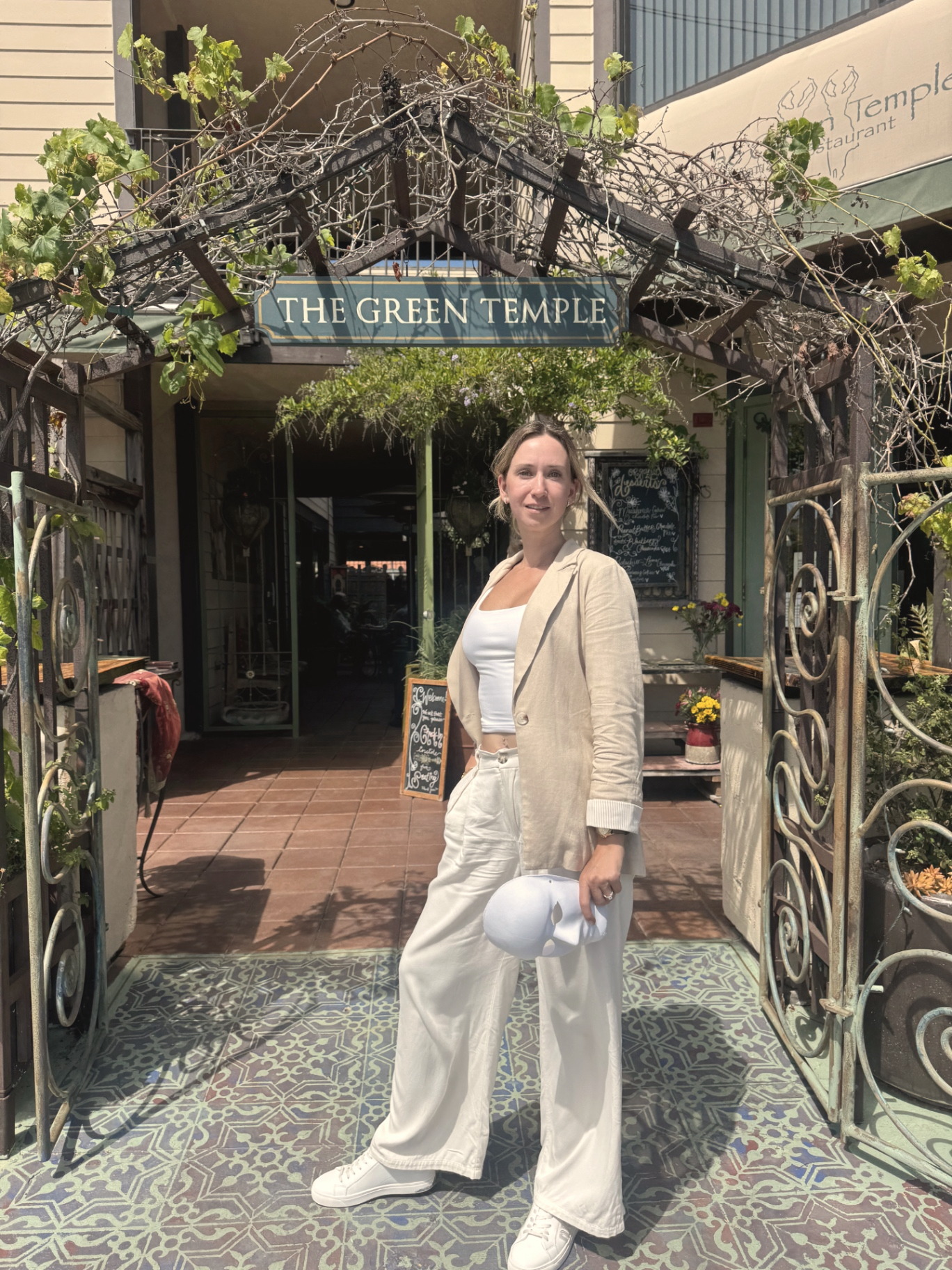Tetiana holding a blank mask at the entrance of a quiet garden restaurant in the South Bay