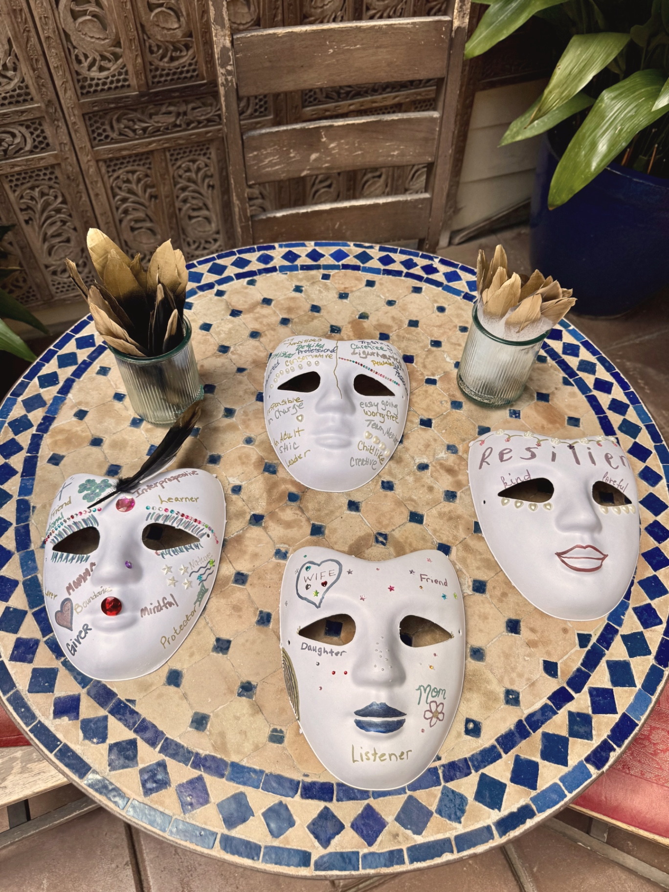 Hand-decorated masks resting on a mosaic table, each one written over with words like mom, wife, listener, resilient